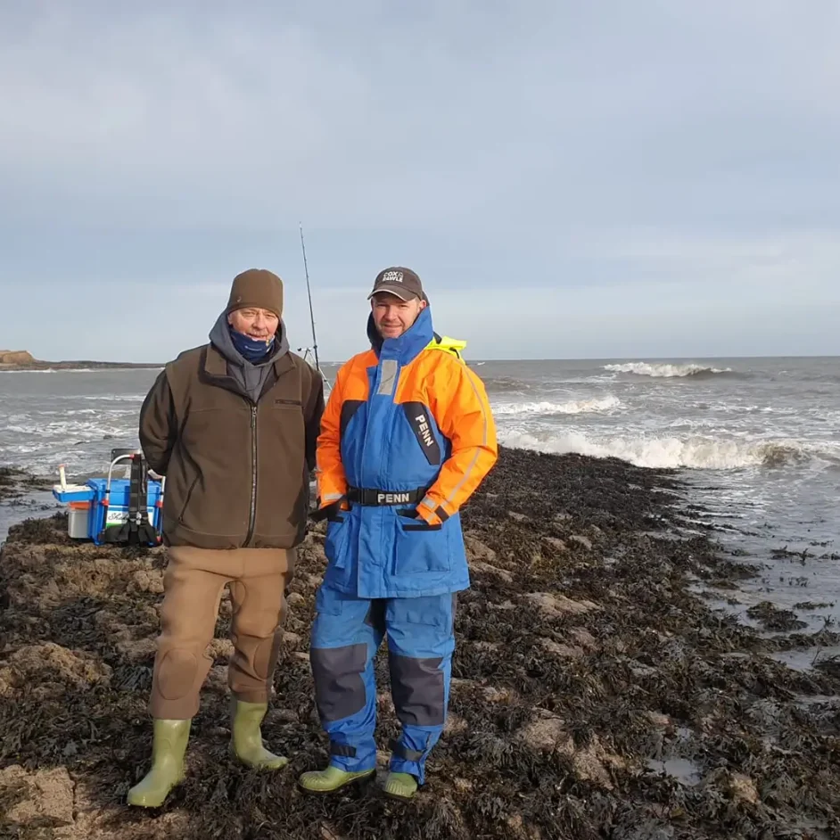 Two fishers stood on coastline during angling activity.